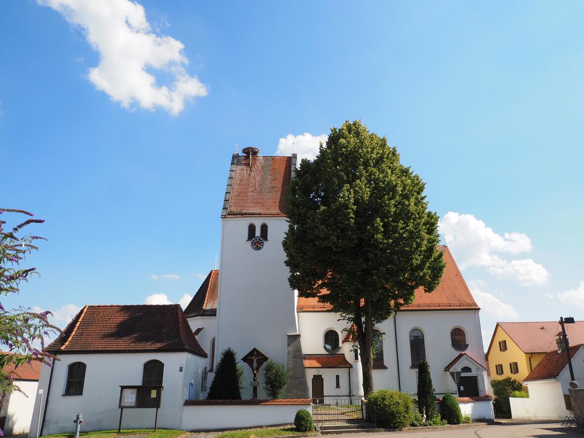 Kirche Hollenbach mit weißer Fassade, spitzem Dach und einem Turm, umgeben von kleinen Bäumen und Sträuchern.