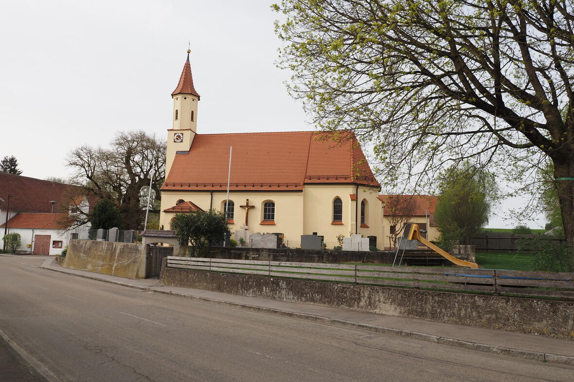 Kirche mit einem roten Ziegeldach, daneben ein Spielplatz und alte Bäume entlang der Straße.