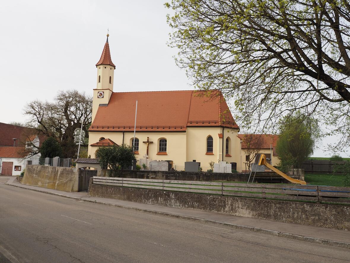 Kirche mit einem roten Ziegeldach, daneben ein Spielplatz und alte Bäume entlang der Straße.